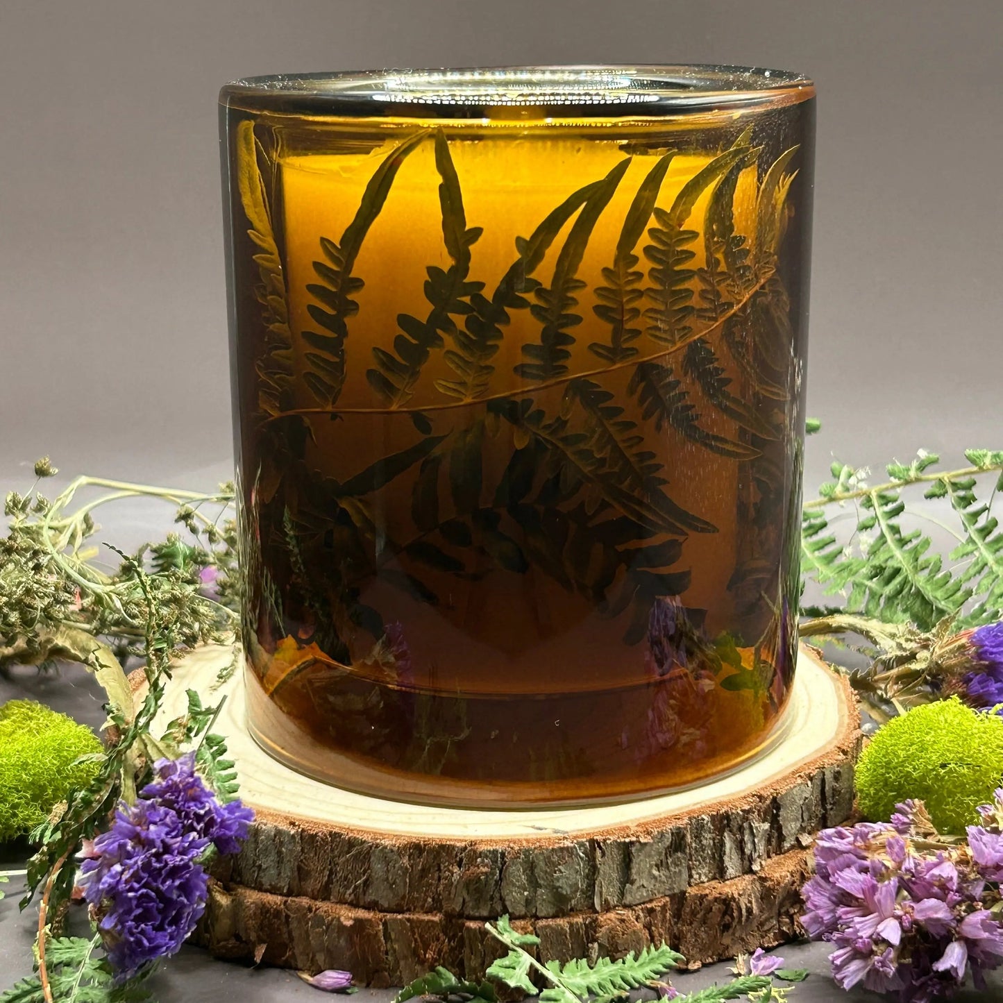 Candle in a see-through orange jar with flowers and greenery placed inside the jar on top of a stack of wood, surrounded by more flowers and greenery.