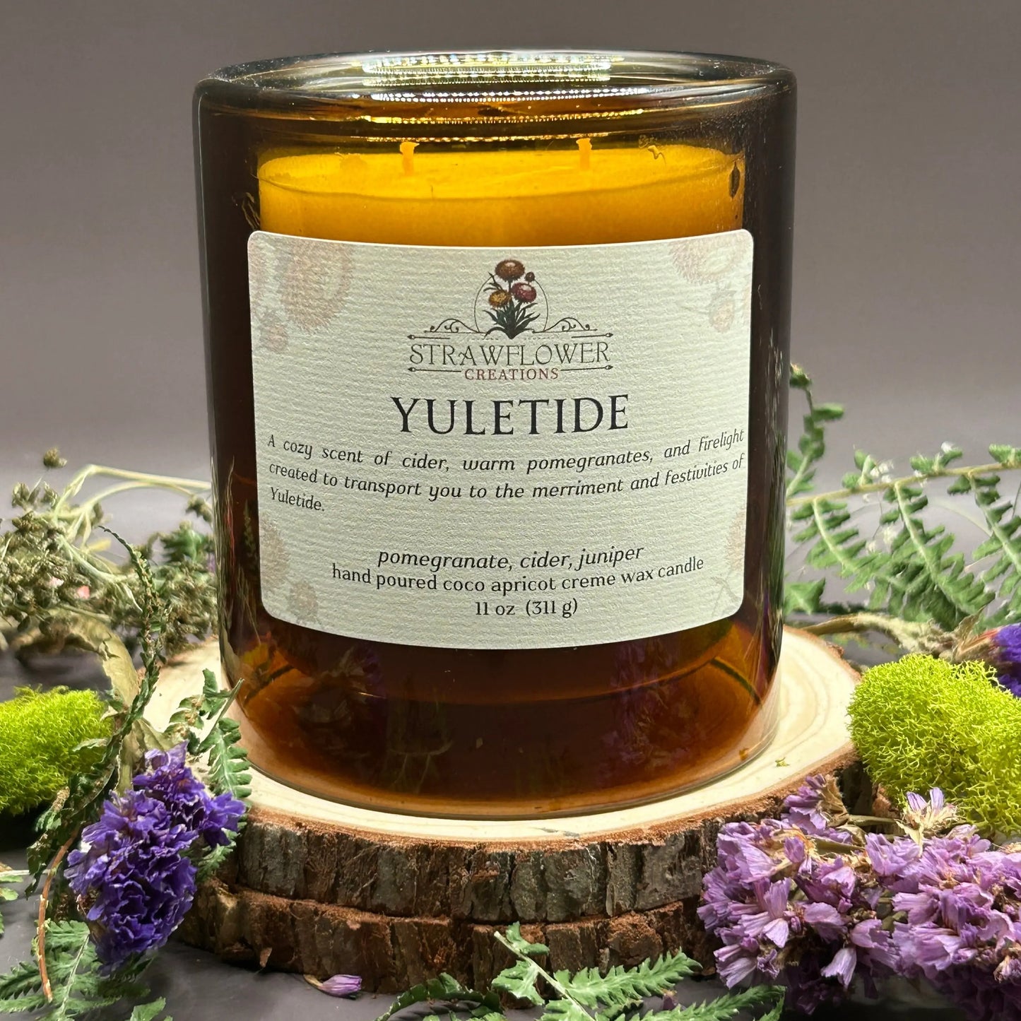 Candle in a see-through orange jar on top of a stack of wood, surrounded by more flowers and greenery