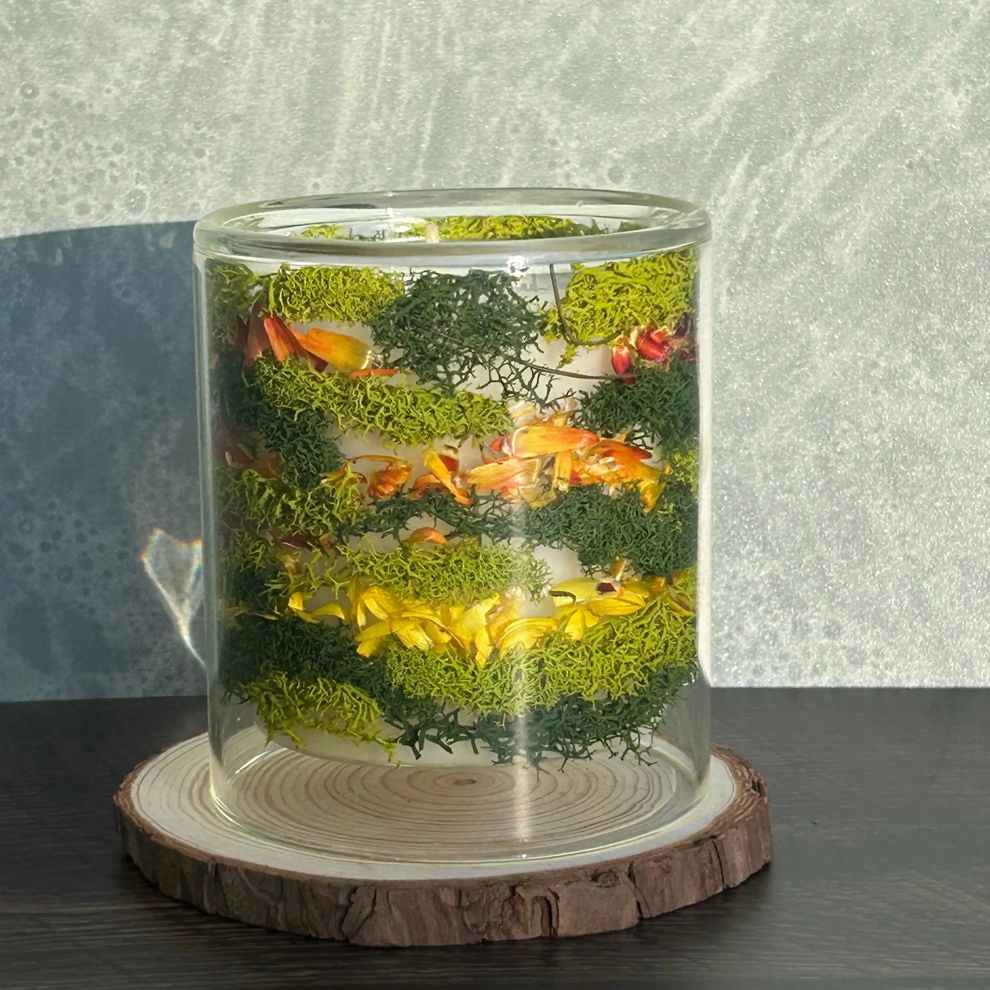 Candle in a clear jar with flowers and greenery placed inside the jar on top of a circle of wood
