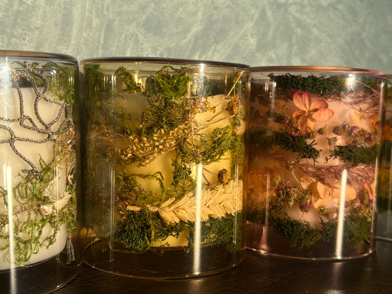 Close up of handmade candles in see-through jars of various colors with flowers and greenery placed in side the jars, sitting on a wooden table with a blue background
