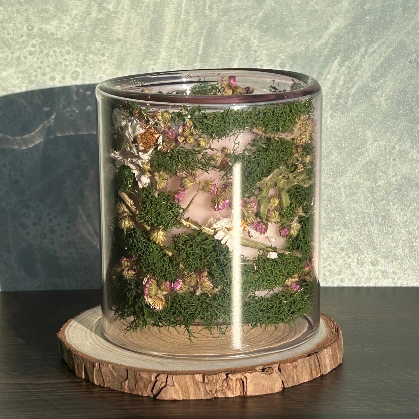 Candle in a clear jar with flowers and greenery placed inside the jar on top of a circle of wood