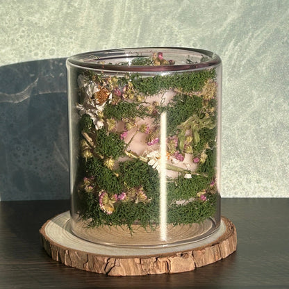 Candle in a clear jar with flowers and greenery placed inside the jar on top of a circle of wood