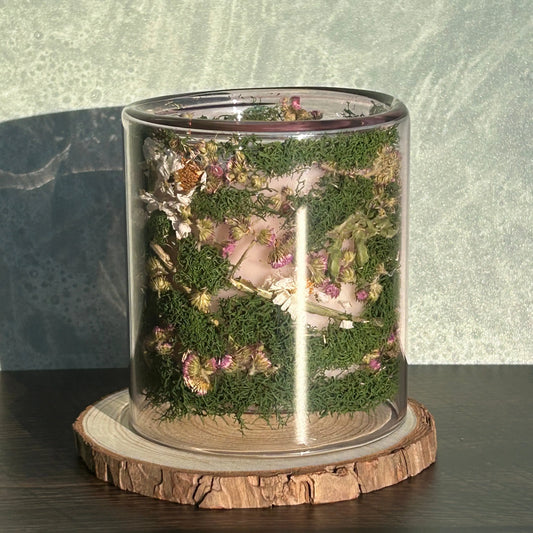 Candle in a clear jar with flowers and greenery placed inside the jar on top of a circle of wood