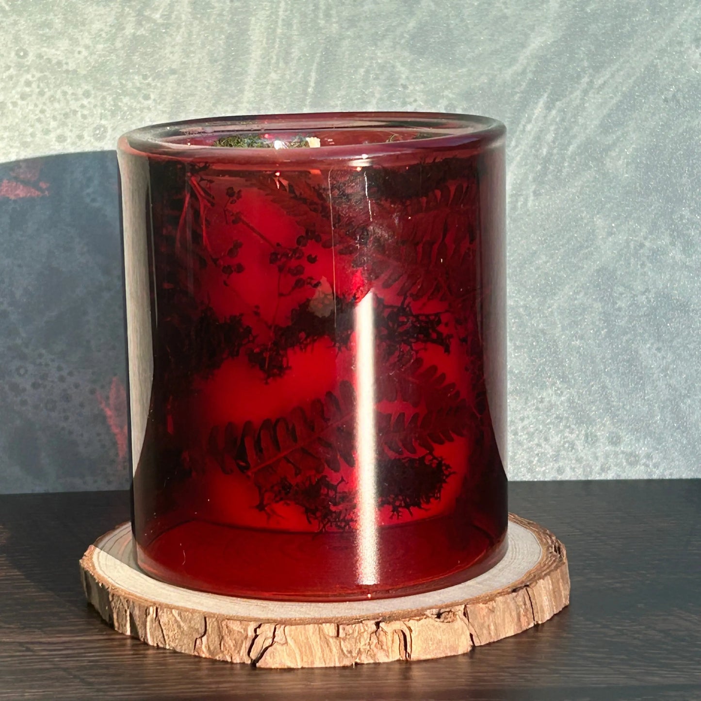 Candle in a see-through red jar on top of a circle of wood.