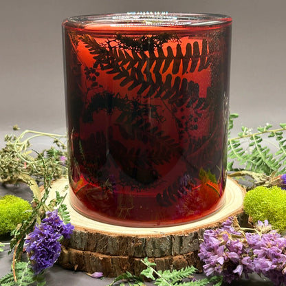 Candle in a see-through red jar with flowers and greenery placed inside the jar on top of a stack of wood, surrounded by more flowers and greenery