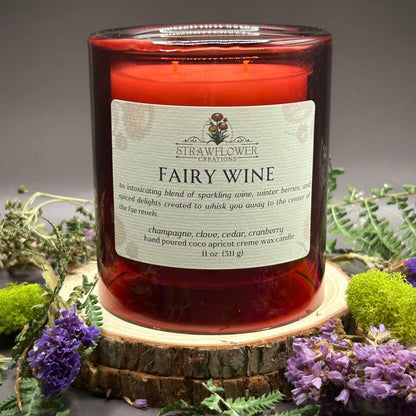 Candle in a see-through red jar on top of a stack of wood, surrounded by more flowers and greenery