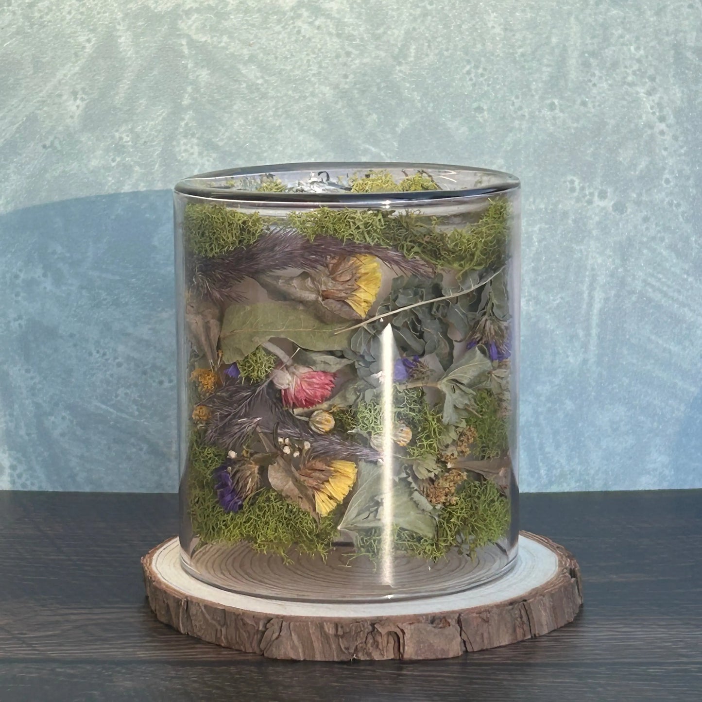 Candle in a clear jar with flowers and greenery placed inside the jar on top of a circle of wood
