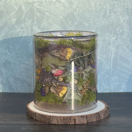 Candle in a clear jar with flowers and greenery placed inside the jar on top of a circle of wood