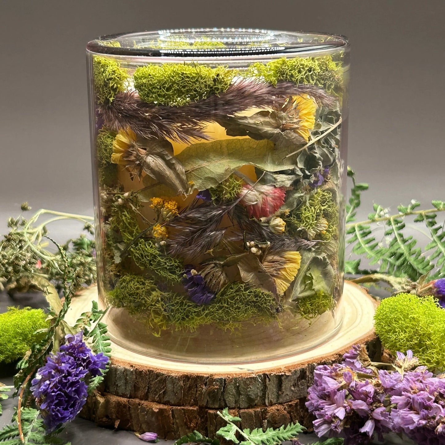 Candle in a clear jar with flowers and greenery placed inside the jar on top of a stack of wood, surrounded by more flowers and greenery