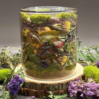 Candle in a clear jar with flowers and greenery placed inside the jar on top of a stack of wood, surrounded by more flowers and greenery