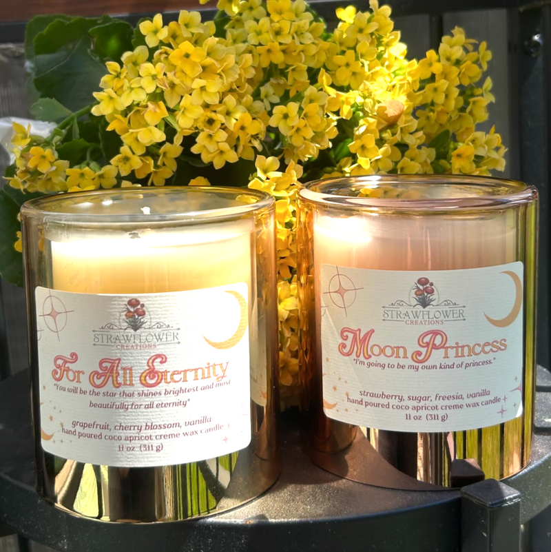 Two candles in see-through jars sit atop a round table in front of yellow flowers