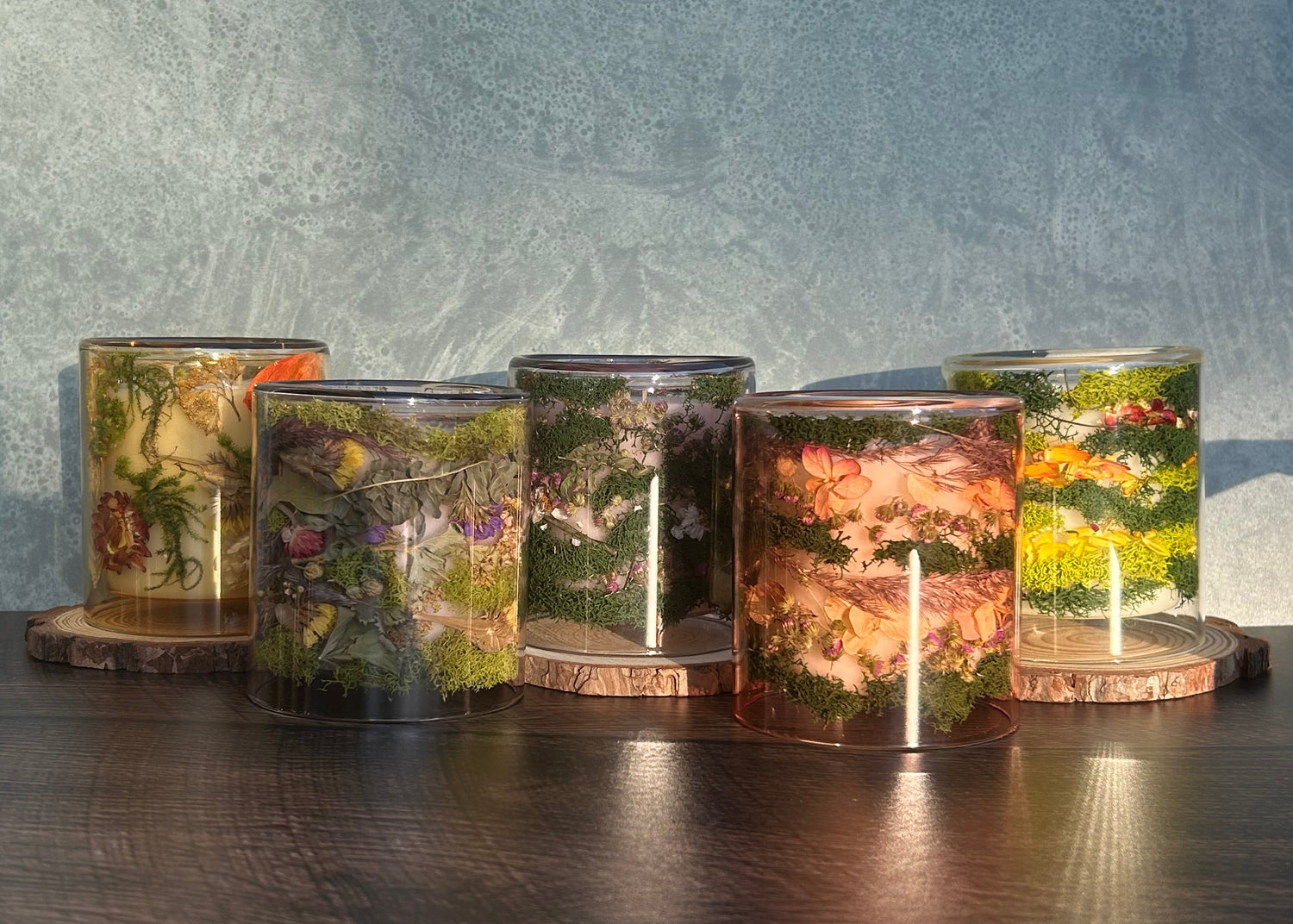 Candles in clear jars lined up on a wooden table with light shining on them onto a blue backdrop