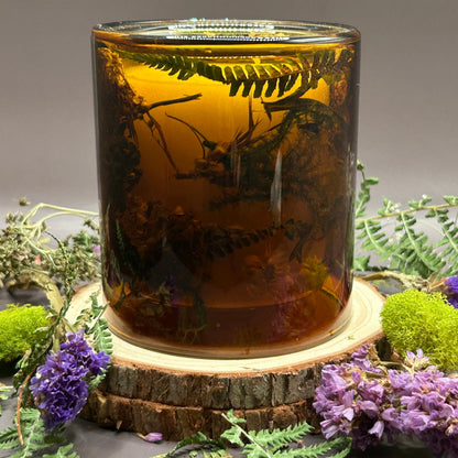 Candle in a see-through orange jar with flowers and greenery placed inside the jar on top of a stack of wood, surrounded by more flowers and greenery