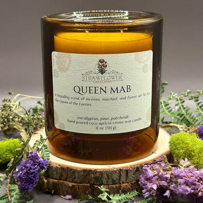 Candle in a see-through orange jar on top of a stack of wood, surrounded by flowers and greenery