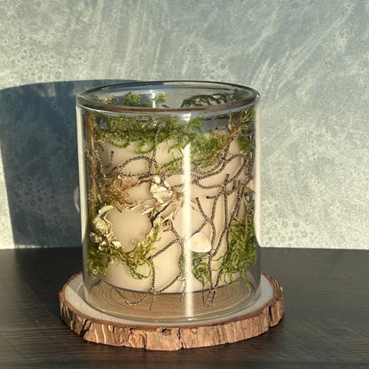 Candle in a clear jar with greenery, rope, and seashells placed inside the jar on top of a circle of wood.