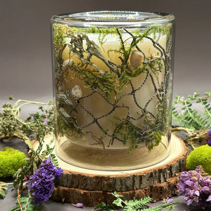 Candle in a clear jar with greenery, rope, and seashells placed inside the jar on top of a stack of wood, surrounded by more flowers and greenery