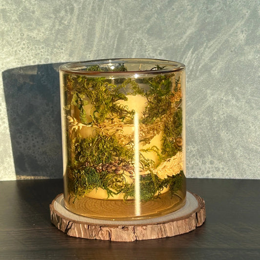 Candle in a see-through yellow jar with flowers and greenery placed inside the jar sitting on top of a stack of wood
