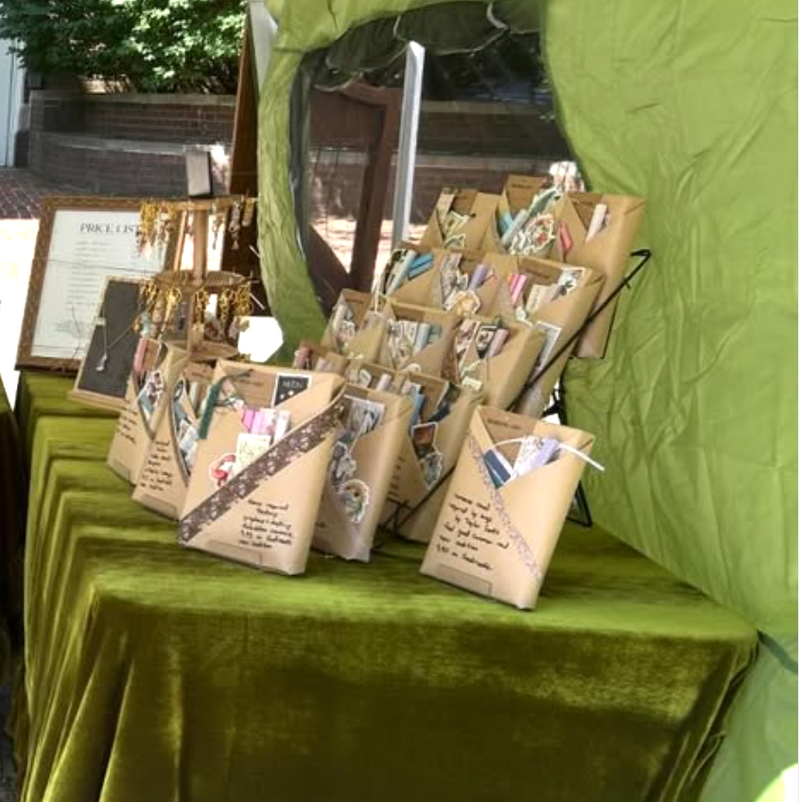 Handmade candles, jewelry, books, and matches line a table covered in a green velvet tablecloth under a tent