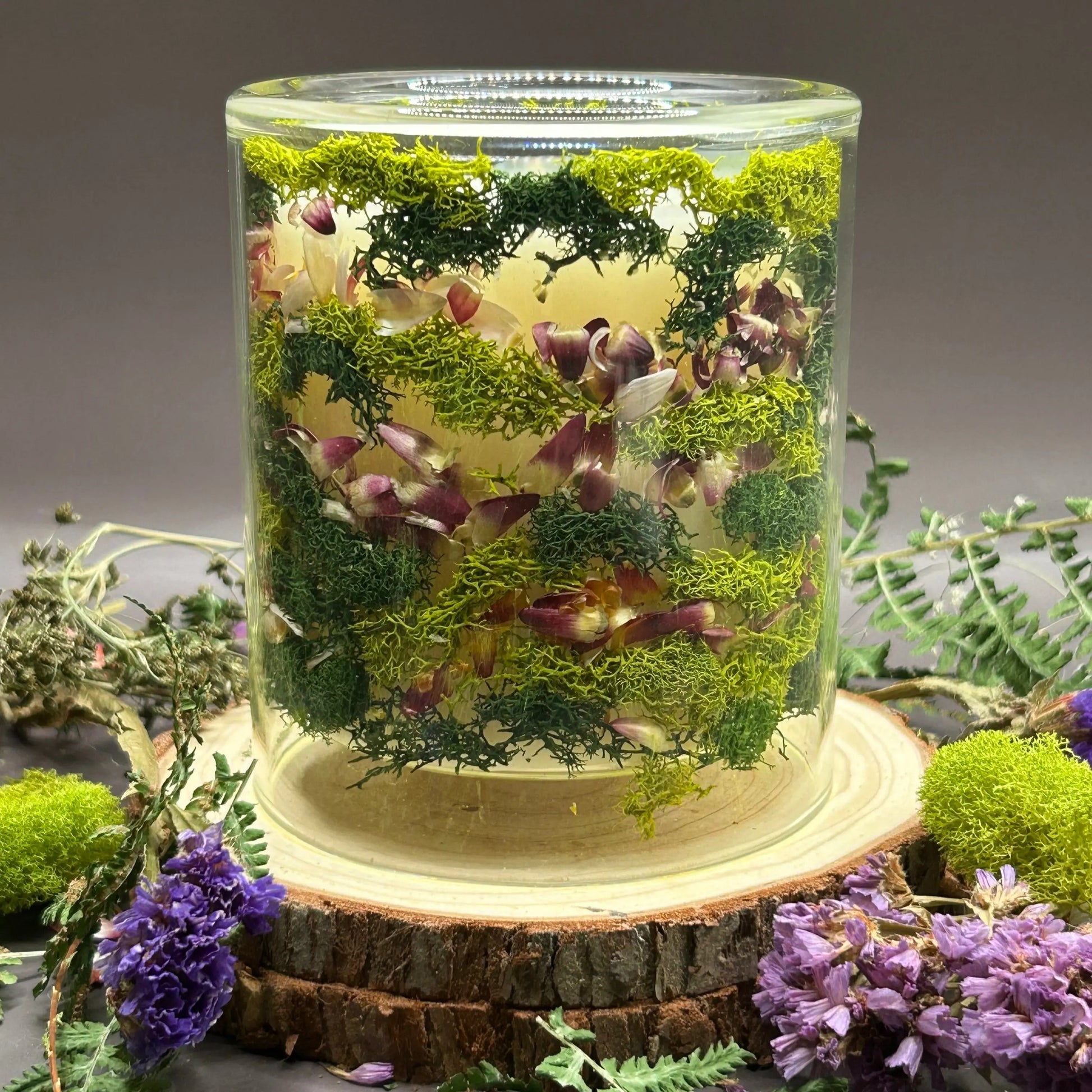 Candle in a clear jar with flowers and greenery placed inside the jar on top of a stack of wood, surrounded by more flowers and greenery