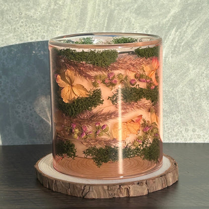 Candle in a see-through pink jar with flowers and greenery placed inside the jar on top of a circle of wood