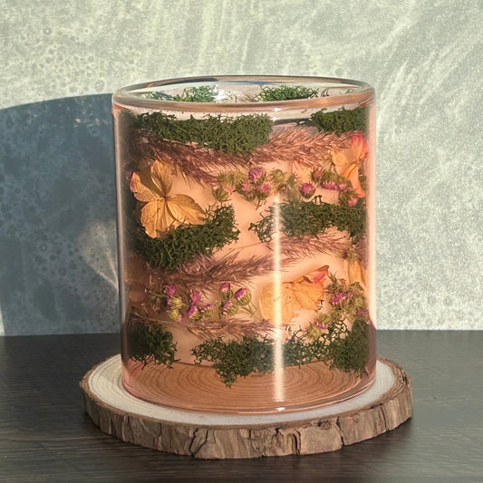 Candle in a see-through pink jar with flowers and greenery placed inside the jar on top of a circle of wood