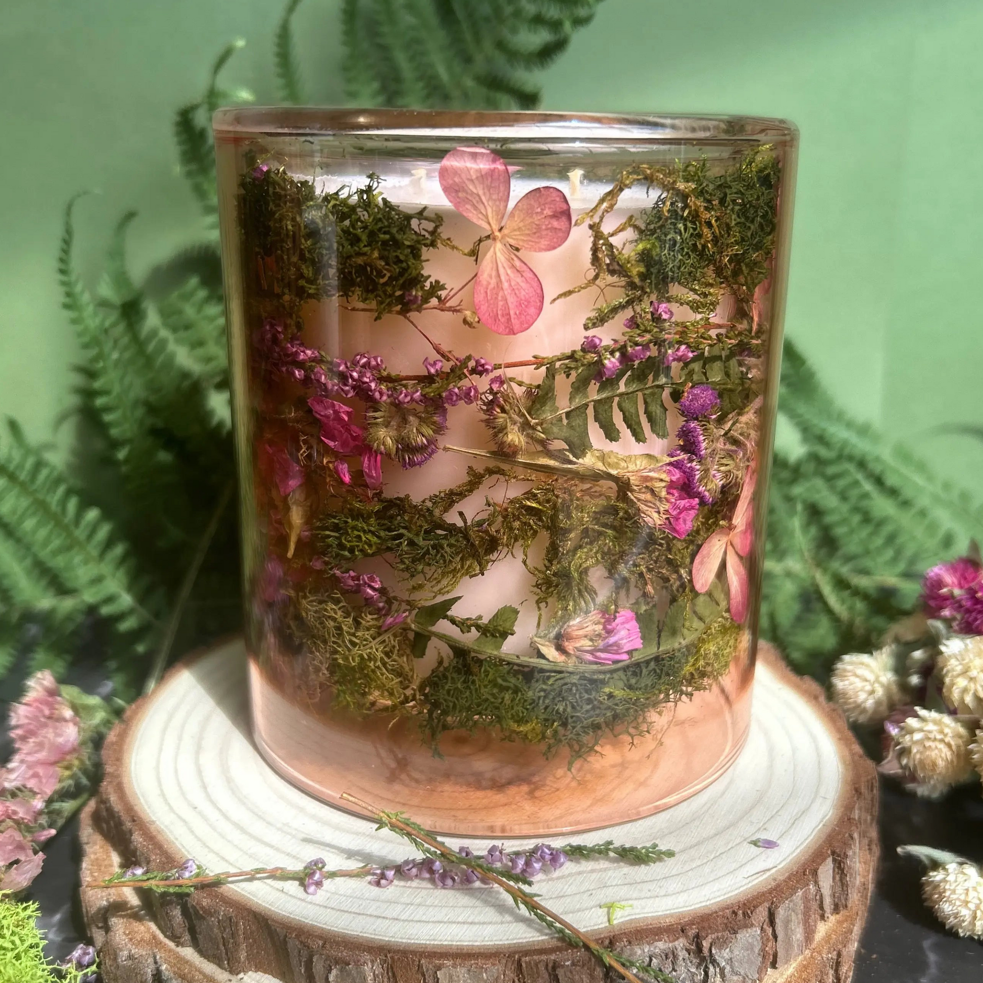 Candle in a see-through pink jar with flowers and greenery placed inside the jar on top of a stack of wood, surrounded by more flowers and greenery
