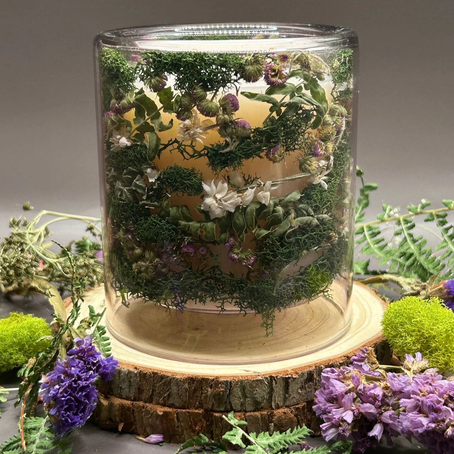 Candle in a clear jar with flowers and greenery placed inside the jar on top of a stack of wood, surrounded by more flowers and greenery