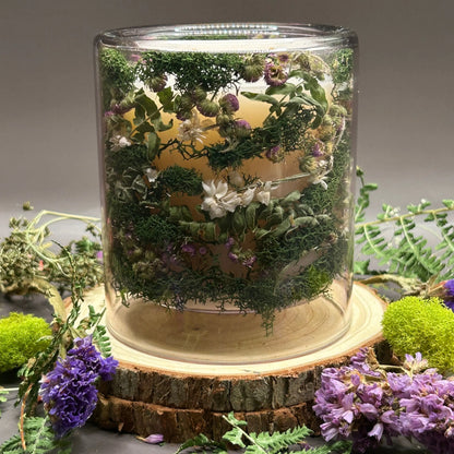 Candle in a clear jar with flowers and greenery placed inside the jar on top of a stack of wood, surrounded by more flowers and greenery