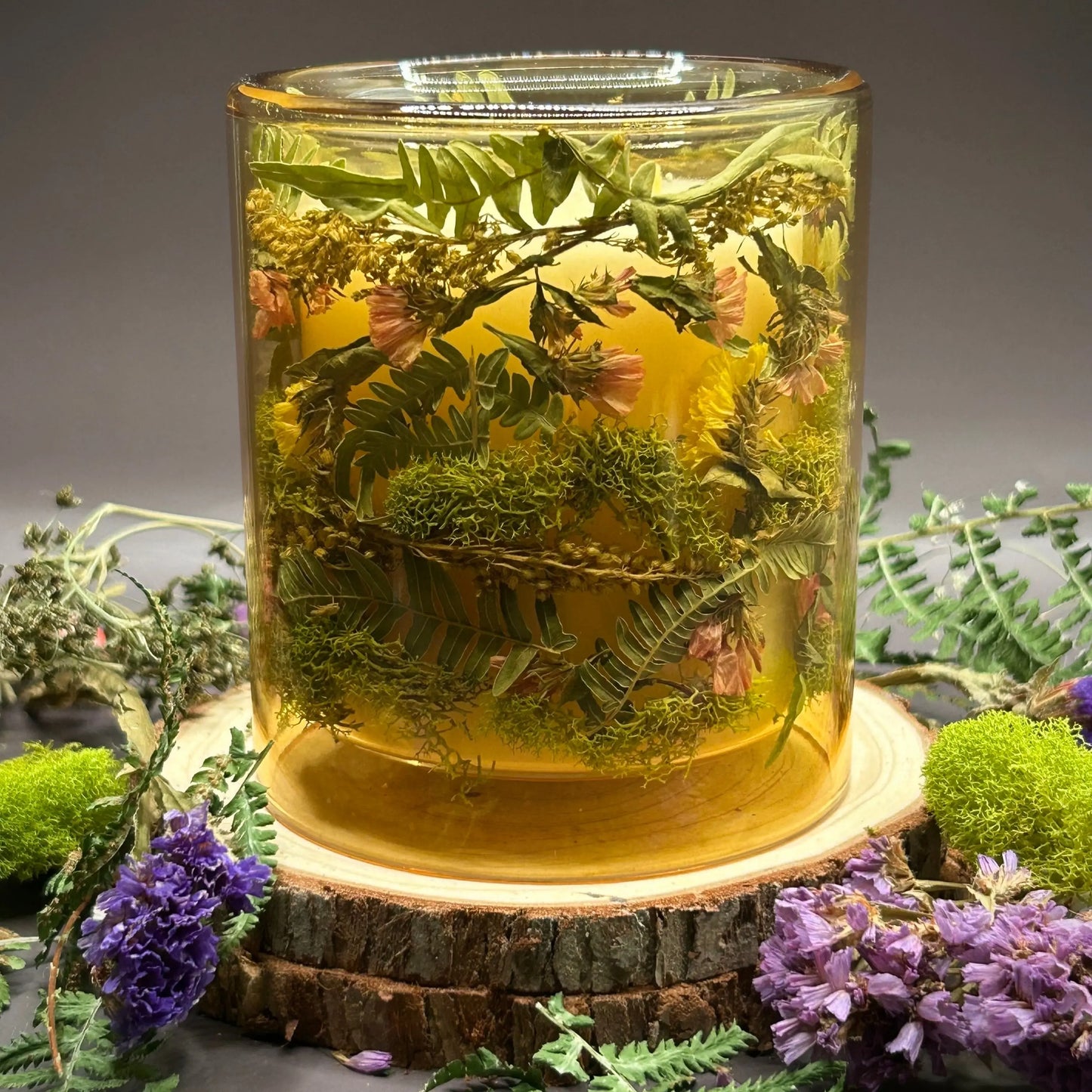 Candle in a see-through yellow jar with flowers and greenery placed inside the jar on top of a stack of wood, surrounded by more flowers and greenery