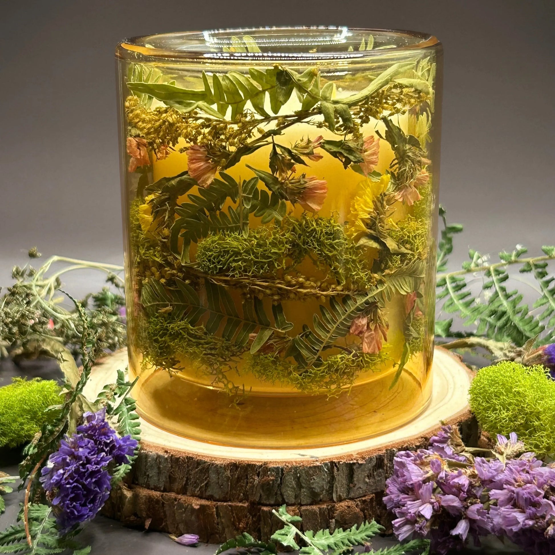 Candle in a see-through yellow jar with flowers and greenery placed inside the jar on top of a stack of wood, surrounded by more flowers and greenery