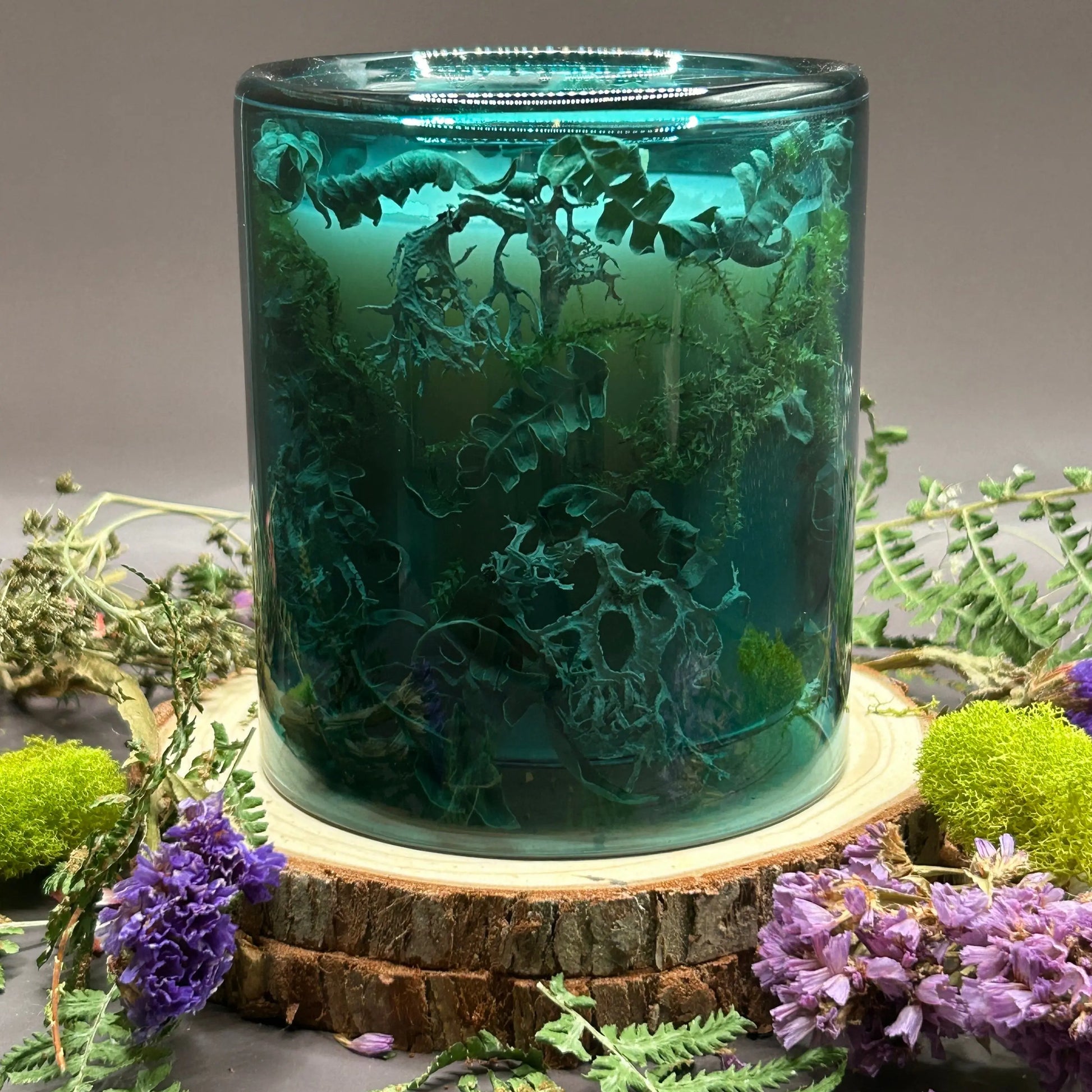 Candle in a see-through green jar with flowers and greenery placed inside the jar on top of a stack of wood, surrounded by more flowers and greenery