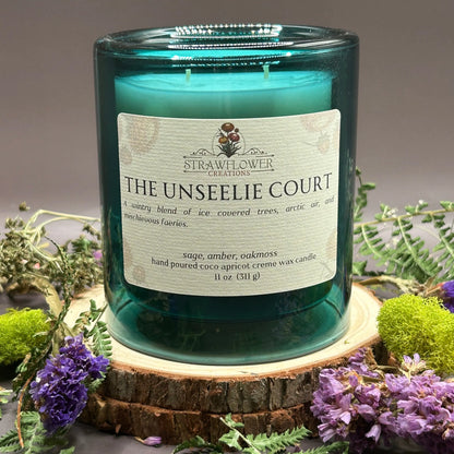 Candle in a see-through blue jar with flowers and greenery placed inside the jar on top of a stack of wood, surrounded by more flowers and greenery