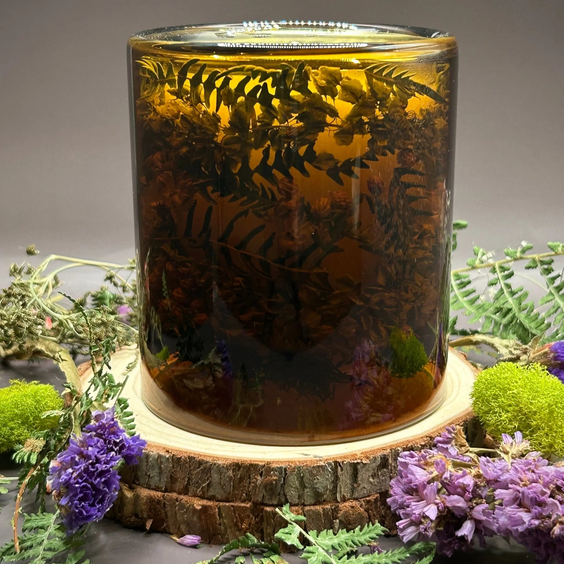 Candle in a see-through yellow jar with flowers and greenery placed inside the jar on top of a stack of wood, surrounded by more flowers and greenery