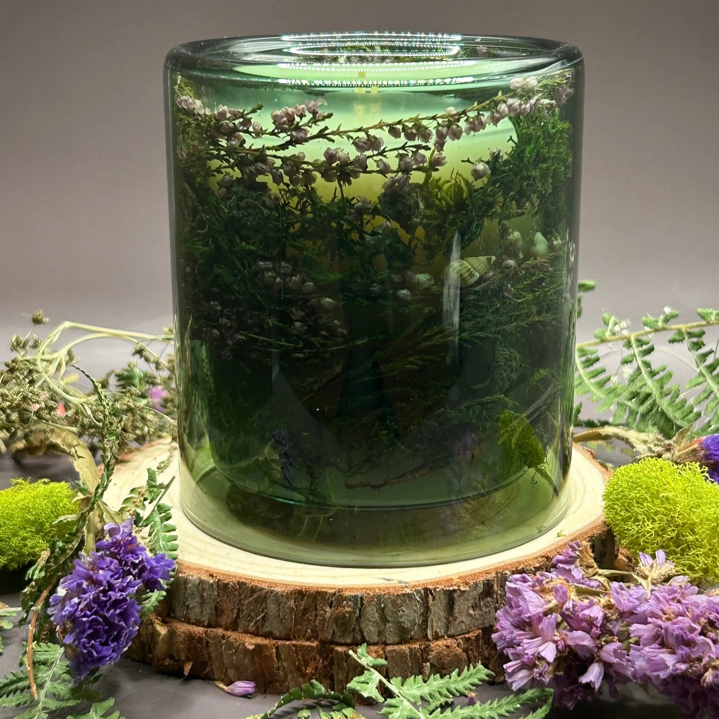 Candle in a see-through green jar with flowers and greenery placed inside the jar on top of a stack of wood, surrounded by more flowers and greenery