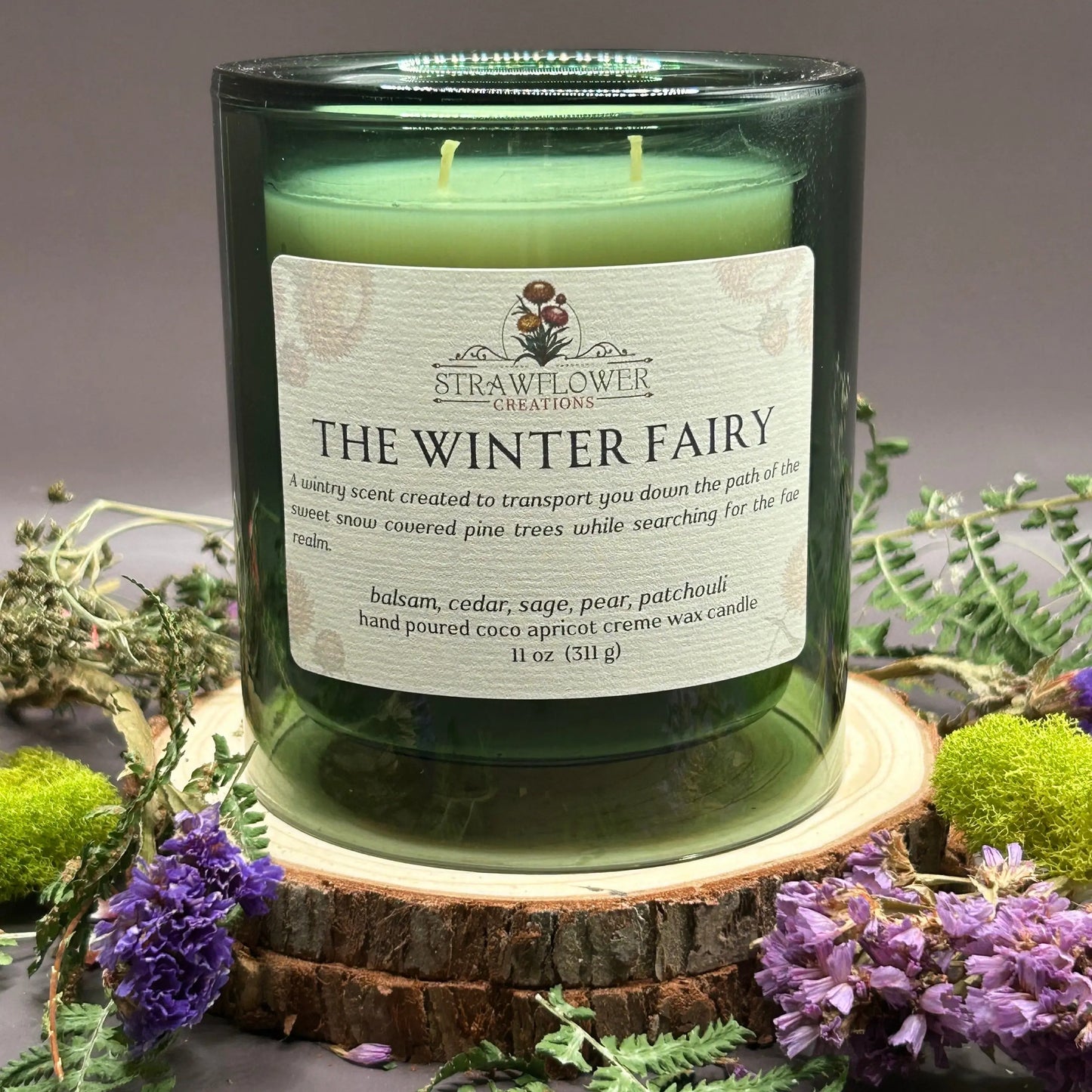 Candle in a see-through green jar on top of a stack of wood, surrounded by more flowers and greenery