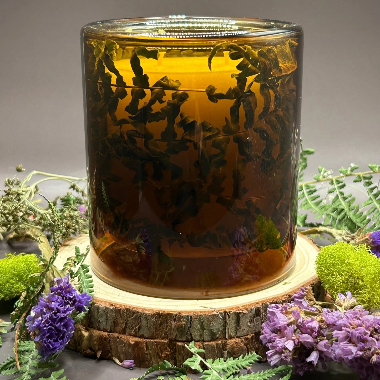 Candle in a see-through orange jar with flowers and greenery placed inside the jar on top of a stack of wood, surrounded by more flowers and greenery