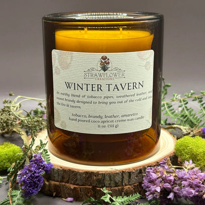 Candle in a see-through orange jar on top of a stack of wood, surrounded by more flowers and greenery