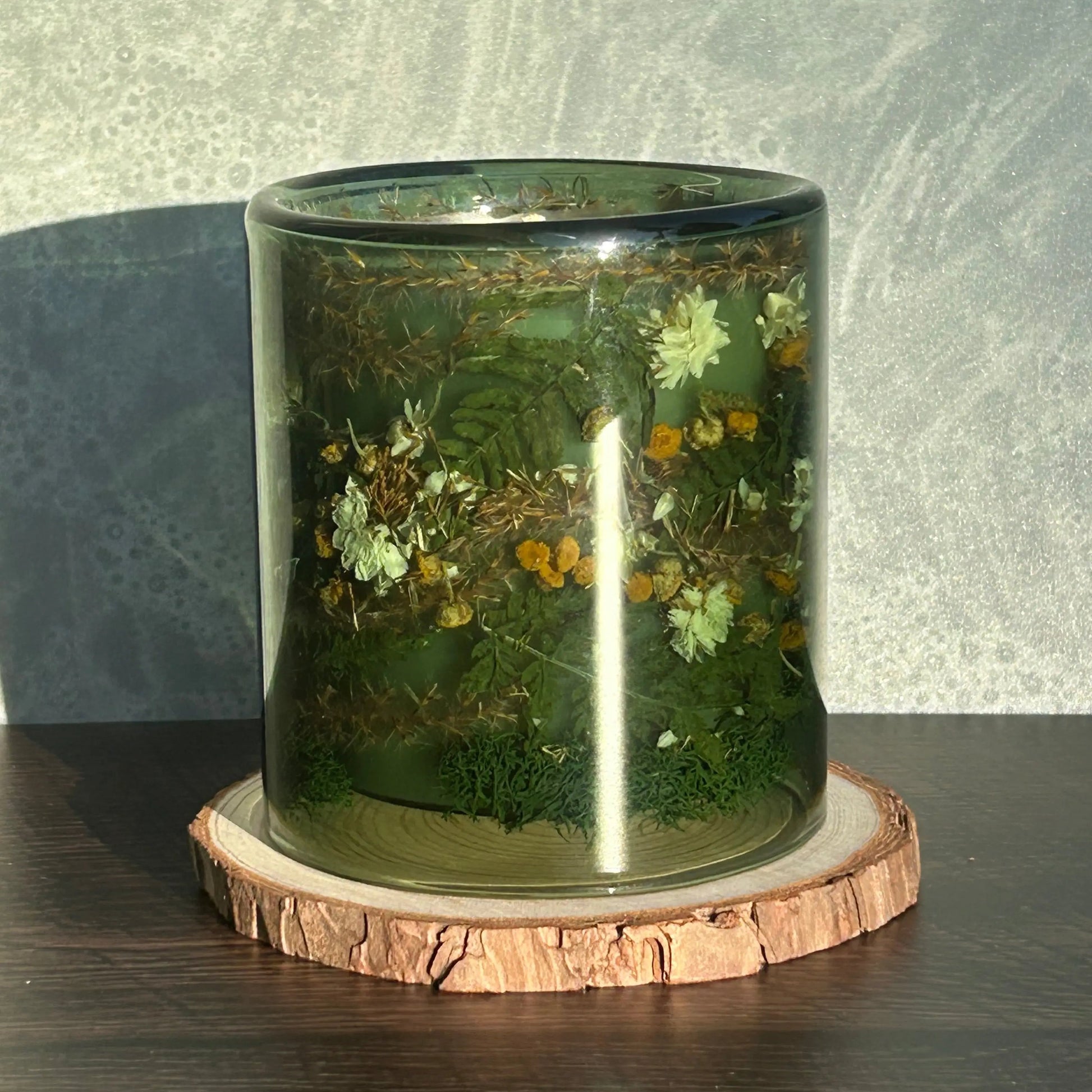 Candle in a see-through green jar with flowers and greenery placed inside the jar, which is sitting on top of a circle of wood. The sun is shining on the jar, creating a glare.
