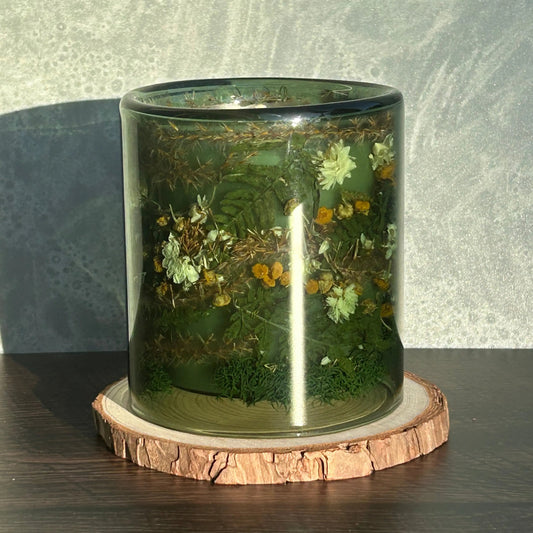 Candle in a see-through green jar with flowers and greenery placed inside the jar, which is sitting on top of a circle of wood. The sun is shining on the jar, creating a glare.