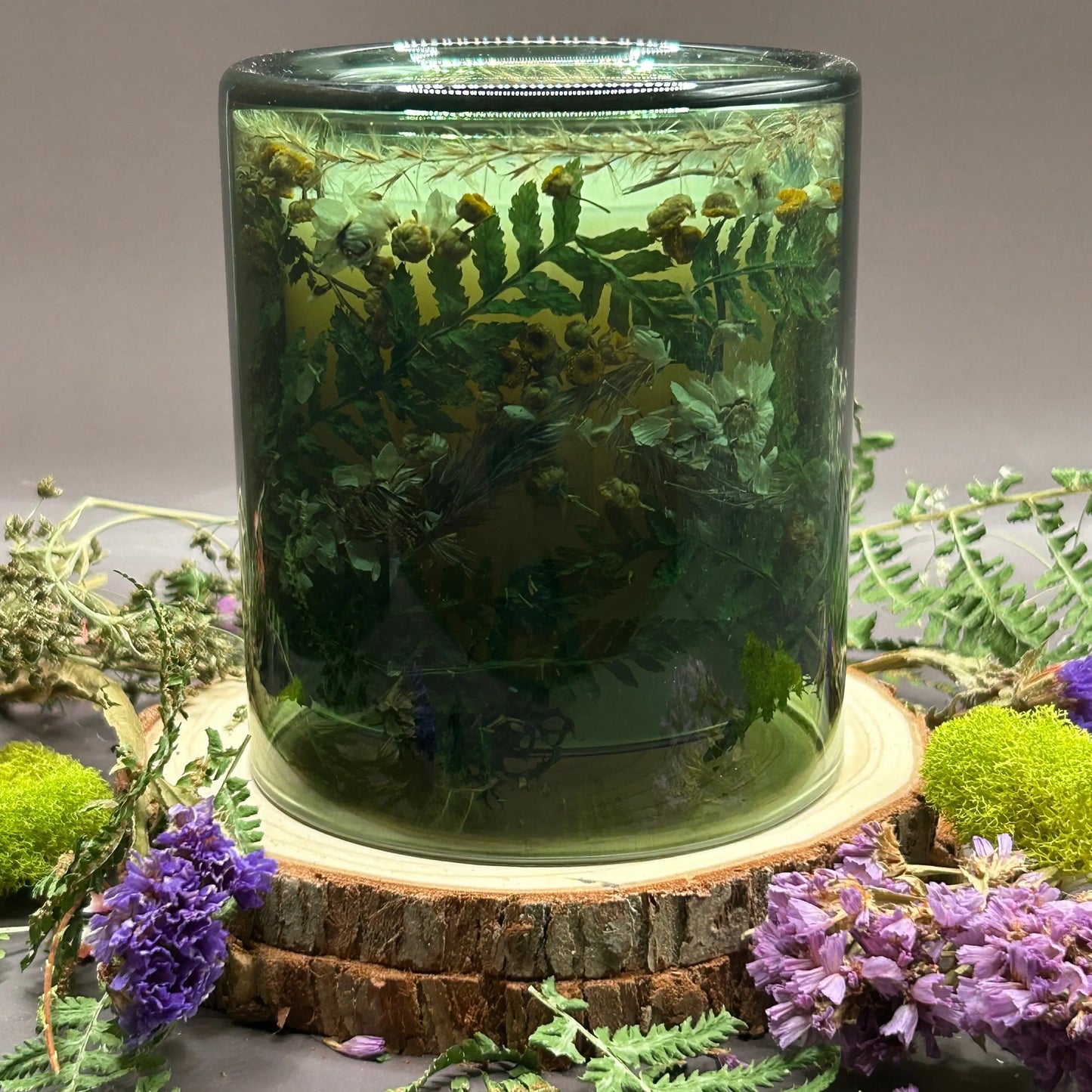 Candle in a see-through green jar with flowers and greenery placed inside the jar on top of a stack of wood, surrounded by more flowers and greenery